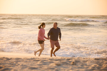 Loving Retired Senior Couple On Vacation Running Along Beach Shoreline Holding Hands At Sunrise