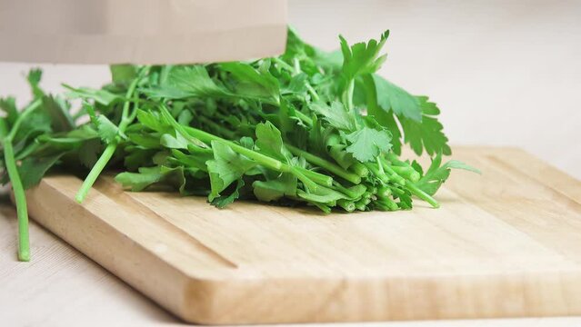 Woman cuts green parsley with a large knife on a wooden board.