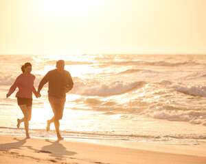 Loving Retired Senior Couple On Vacation Running Along Beach Shoreline Holding Hands At Sunrise - 763983498