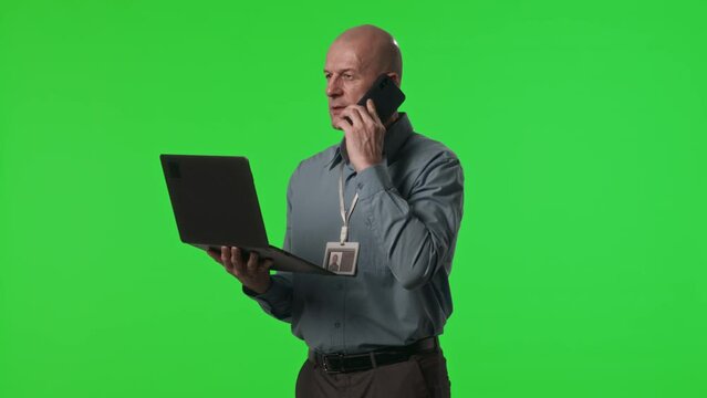 Mid-aged data center engineer holding laptop and talking on mobile phone in studio with green chroma key background