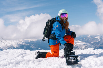 woman snowboarder fastens snowboard bindings while standing on top of  slope in ski resort. Against background of grandiosely beautiful mountain range - obrazy, fototapety, plakaty