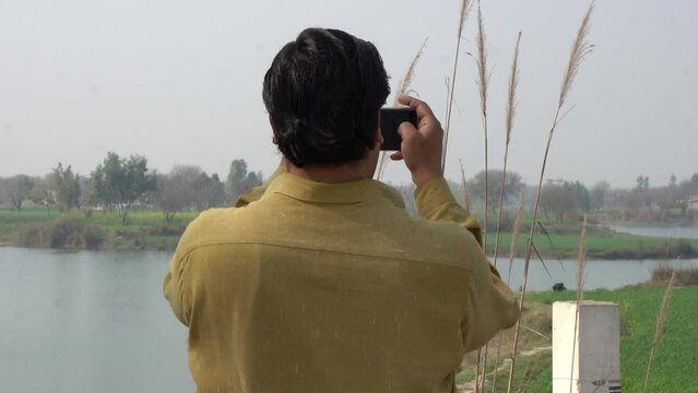 A man is standing on the bank of the river making a video with his mobile phone. Young hipster guy taking photo on his mobile phone camera of a beautiful landscape while standing near lake.Slow Motion