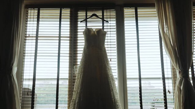 A wedding dress hangs on a wooden hanger by the window