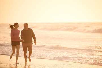 Fototapeta na wymiar Loving Retired Senior Couple On Vacation Running Along Beach Shoreline Holding Hands At Sunrise