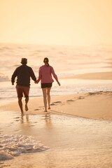 Rear View Of Retired Senior Couple On Vacation Running On Beach Shoreline Holding Hands At Sunrise