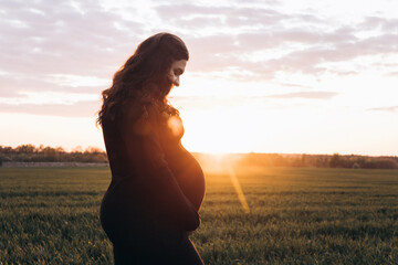 A pregnant woman is holding her stomach in the middle of a field. A girl is expecting a baby at sunset in summer. Fatherhood and motherhood among young people - obrazy, fototapety, plakaty
