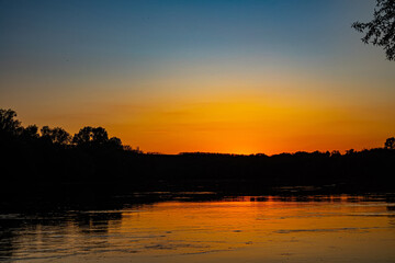 Great Don River in middle reaches. Orange sunset is reflected in water. Black silhouette of coastal forest. There are many trichopterans flying over smooth whirlpool current. Spring and high water