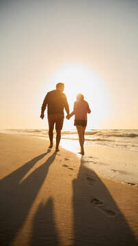 Rear View Of Retired Senior Couple On Vacation Walking On Beach Shoreline Holding Hands At Sunrise