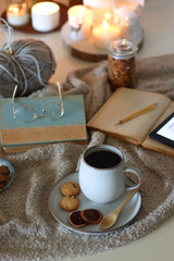Cup of tea or coffee, cookies, books, glasses, e-reader, almonds, ball of yarn, knitting needles and lit candles on the table. Hygge at home. Selective focus.