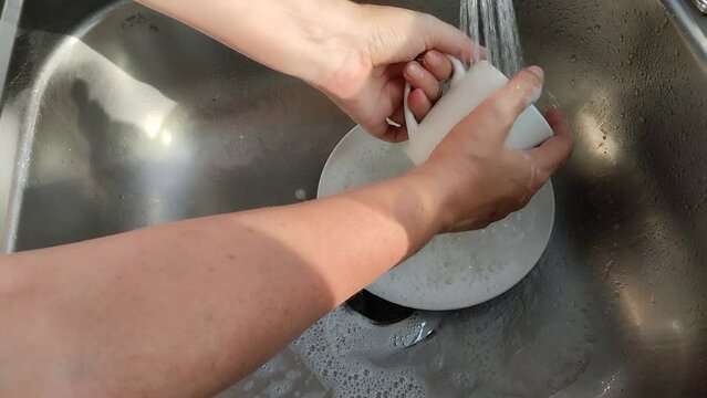 Unas manos de  mujer lavando una  taza y un plato de color blanco  con  agua  en un fregadero