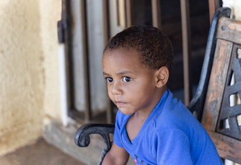 african boy with a serious look, sited on a bench, in front of the house in the township, late...