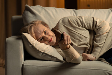 A mature woman lies comfortably on a couch, her body draped over soft cushions, as she holds a...