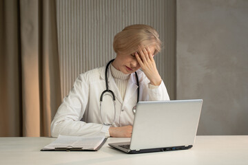 A weary doctor, stethoscope around her neck, sits at a desk with her hand on her forehead,...