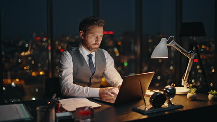 Late businessman searching data typing laptop at night cityscape in office.