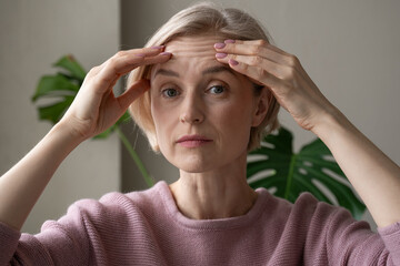A contemplative mature woman is captured indoors while gently touching her forehead with both...