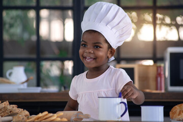 Happy little African girl wear chef hat in bakery kitchen, she learned about baking, smiling at...