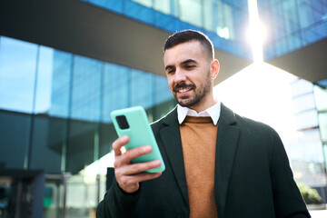 A young Caucasian man in a formal suit standing in front outside an office building using and...