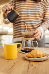 female makes coffee in morning kitchen. brews in purover. yellow cup with black coffee drip. daily routine of happy woman. Food and drinks, croissant on table. Americano cappuccino for cheerfulness