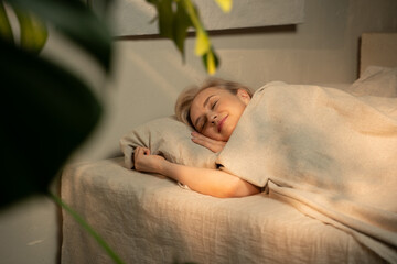 A woman is captured in a serene slumber, nestled under a light blanket in a warmly lit, inviting bedroom setting. The room exudes a tranquil atmosphere, suggesting a late evening or nighttime