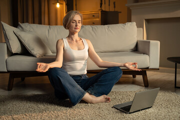 A serene mature woman sits cross-legged on a cozy living room floor, engaging in a meditation...
