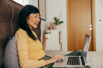 A happy japanese woman is sitting on a bed, drinking coffee and scrolling on a laptop. - Powered by Adobe