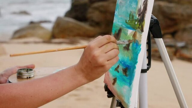 Artist paints seascape on sandy shore. Creative female uses brush, palette for art piece. Canvas captures ocean waves, coastal scene. Outdoor inspiration, leisure activity by water. Slow motion.