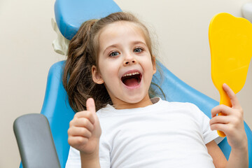 Portrait of a cute happy little girl sitting on a chair in a dental office, showing thumbs up after a dental examination while holding mirror in her hand.