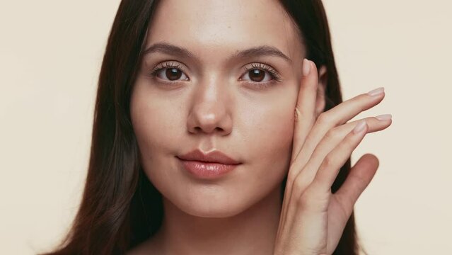 Calm young woman with a serene expression and her hand gently resting on her face against a neutral beige backdrop.