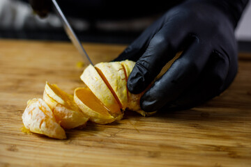 Person with black gloves cuts peeled orange on wooden cutting board into pieces