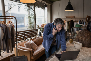 Hispanic male using a laptop and phone computer in his small business vintage clothing store