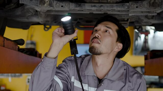 Young asian mechanic with light in hands looking at steering chassis and suspension under a car of a lifted car, Garage service. Automobile diagnostic, Car service and auto repair concept
