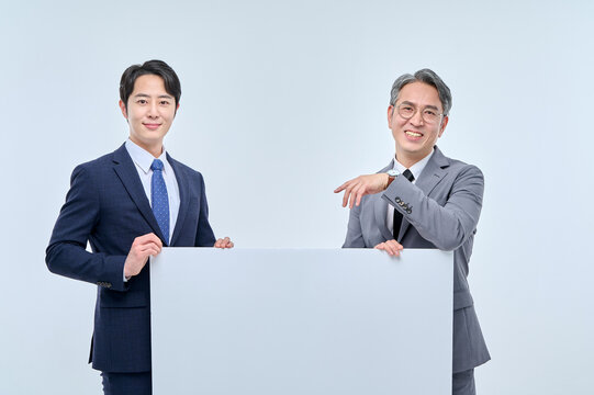 A young office worker man wearing a suit and glasses and a middle-aged man are holding a white board together and posing with various confident expressions and poses.