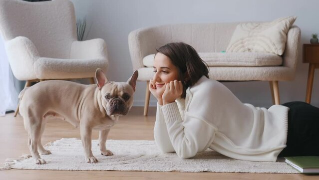 Looking at each other, lying down on floor. Young woman is with her pug dog at home.