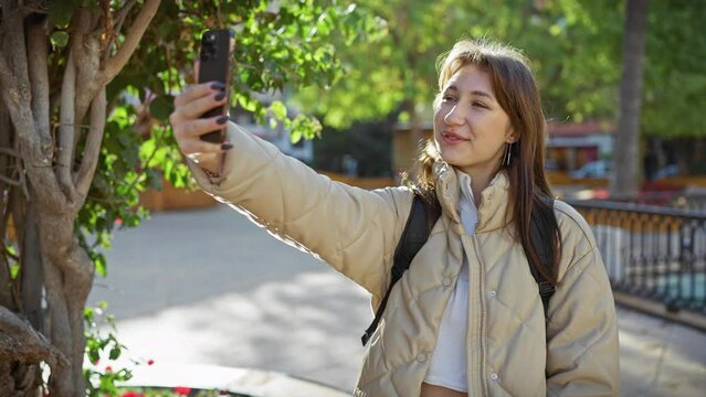 A young woman takes a selfie outdoors in a park with greenery, using a smartphone and wearing a casual jacket.