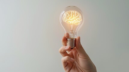 Hand holding a light bulb with a white bright glowing brain inside against gray background.