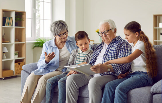 Portrait of a happy grandparents reading a book to their grandchildren or looking through a family photo album sitting on sofa at home. Kids enjoying leisure time with grandma and grandpa on weekend