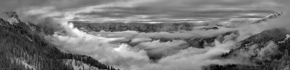 Winter in the mountains: black and white processed large Panorama of the Pale di San Martino valley, view from the air. Dramatic cloud cover, snow covered trees and surroundings. Italy, Dolomites.