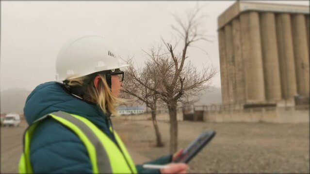 Girl student in hardhat and green reflective vest recording notes on tablet, standing at construction site while learning professional skills during an internship in remote factory area. 