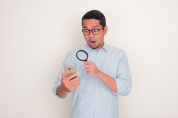 Adult man showing shocked expression when looking mobile phone using magnifying glass