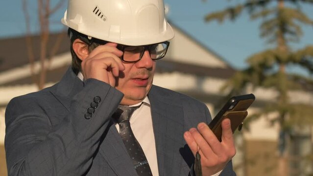 Strict boss of construction company types message on touchscreen with finger in phone, adjusts glasses standing in white protective helmet, suit and ties, close-up on country houses background.