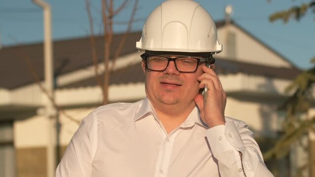 Close-up of handsome mature man i strict white shirt in a protective helmet with glasses holding phone near ear, talking and gesturing with hands on street during day.