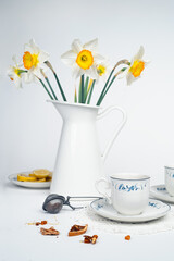 Still life with a fine porcelain tea cups and accessories arranged with a blooming bouquet of white daffodils on a textured white background - 764139243
