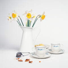 Still life with a fine porcelain tea cups and accessories arranged with a blooming bouquet of white daffodils on a textured white background - 764139406