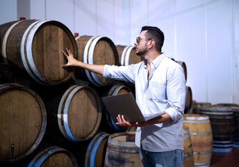 Professional man winemaker or owner working and inspecting wine quality in wine cellar with wooden barrel in wine factory. Winery manufacturing industry, Alcohol and winemaking fermentation process.