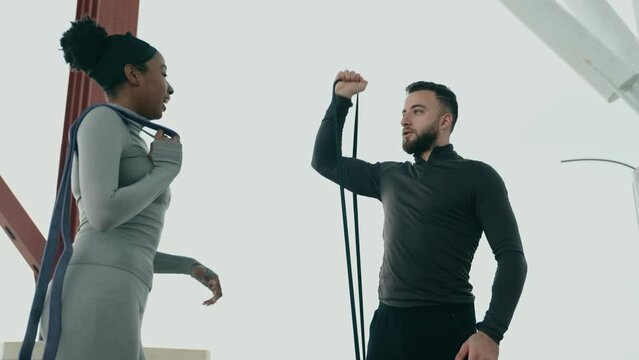 Multiethnic fitness duo during an outdoor training session. A Middle Eastern man exercises with resistance bands while a Black woman observes and advises.