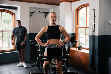 A woman is cycling on an exercise bike with a man standing behind her at the gym