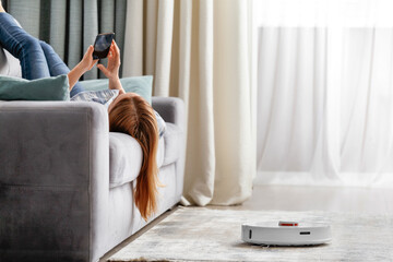 The girl lies on the sofa in the living room and controls a smart robot vacuum cleaner using a...