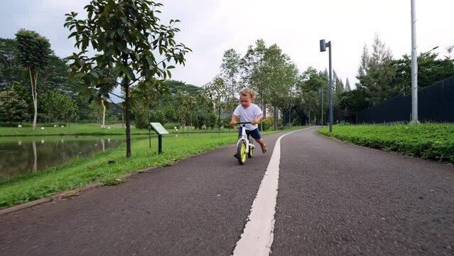 Spirited young child races through verdant park on balance bike, confidently propelling himself forward with each push of his feet. Scene captured in slow motion with reverse dolly shot