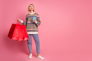 Full size photo of cheerful girl wear ornament sweater hold shopping bags smartphone look at empty space isolated on pink color background