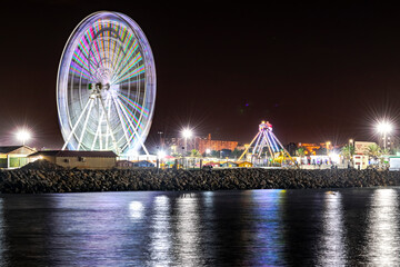 Ferris wheel and pirate ship in the amusement park seen from Sablettes beach promenade in the dark. Water surface view of Algiers city by night, water reflexion of electric lights in the rock jetty.
